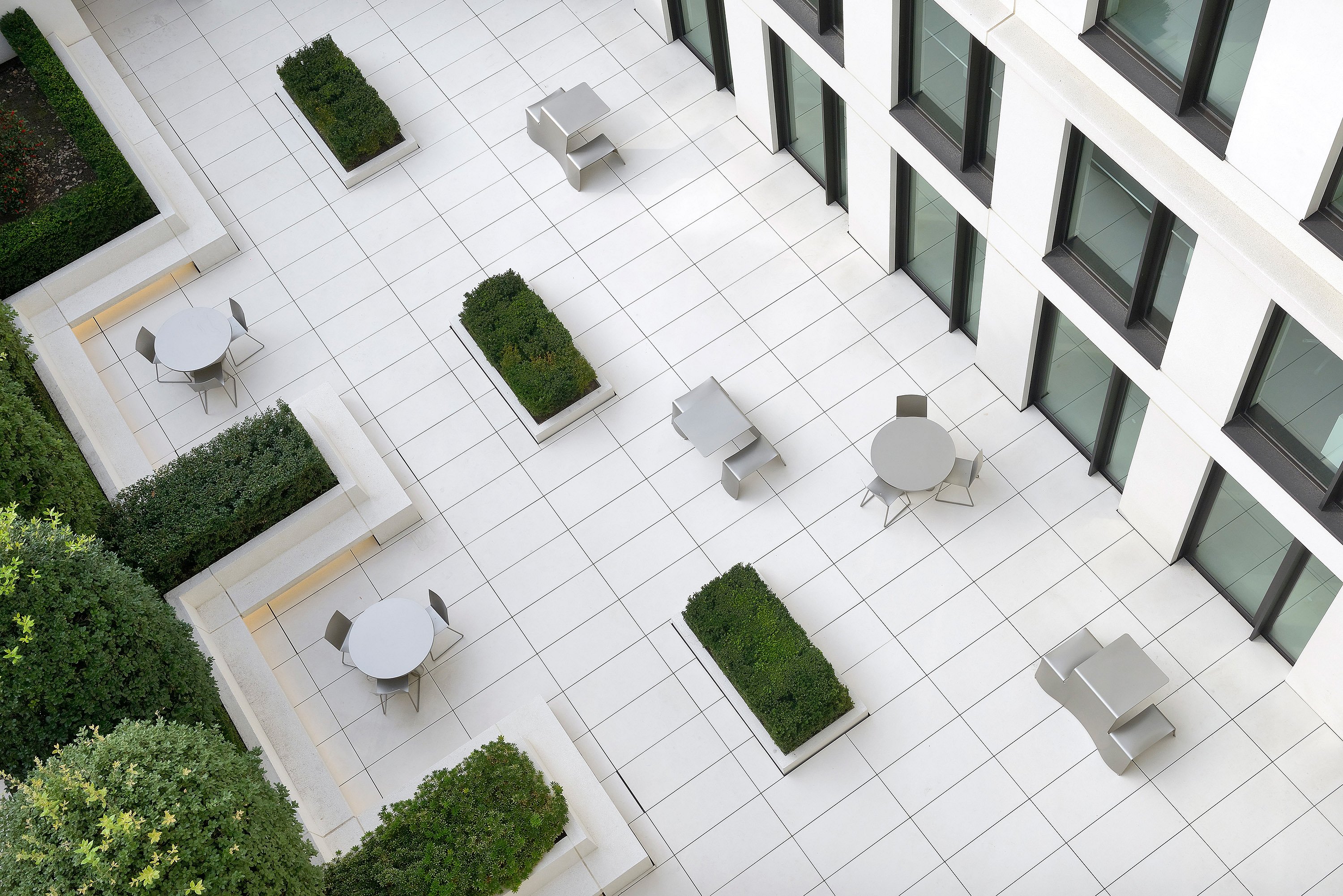 Blick von oben auf einen strahlend weißen Bürohof mit runden Tischen, rechteckigen Pflanzgefäßen und blassen, skulpturalen Picknickbänken, die zwischen den Fassaden des Gebäudes angeordnet sind.