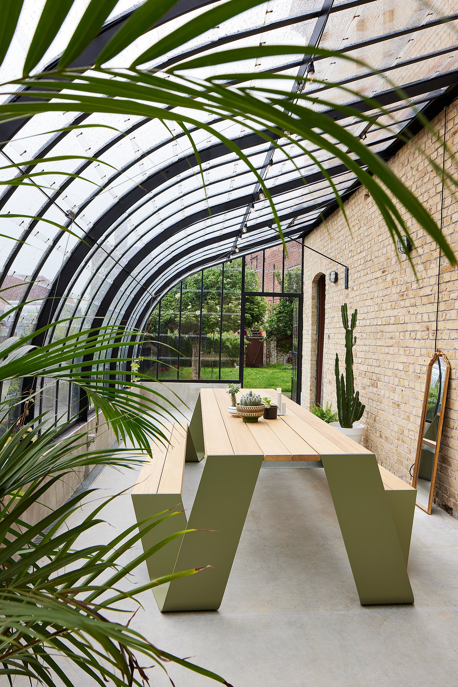 Een groene Hopper houten picknicktafel in een serre met glazen dak en tropische planten.