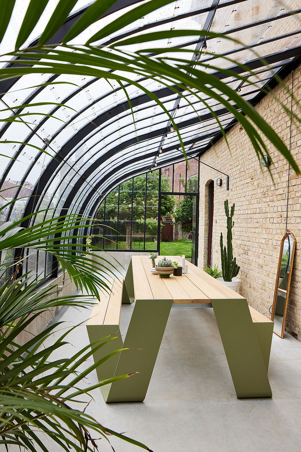 Leeres Gewächshaus-Interieur mit einem langen hölzernen Trichter-Picknicktisch mit grünen Beinen in der Mitte auf einem glatten Betonboden, eingerahmt von Palmblättern, Ziegelwänden und schwarzer Stahlverglasung.