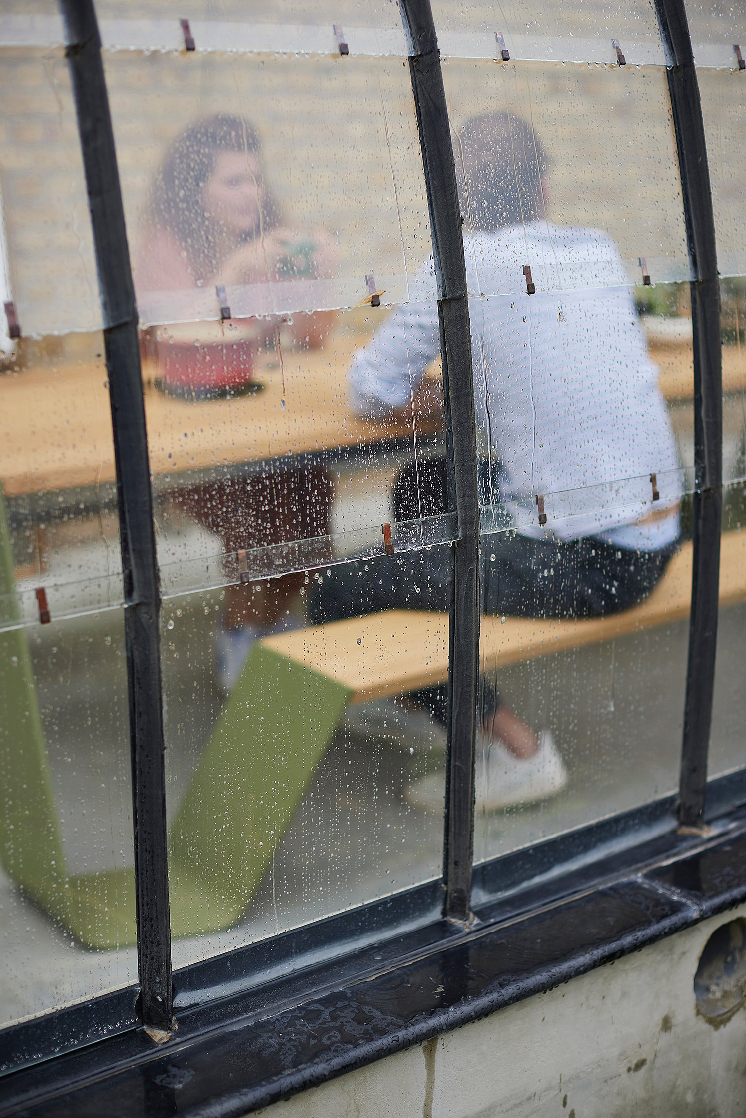 Verschwommener Blick durch regengepunktete Gewächshauspaneele von zwei Personen, die am Trichterpicknicktisch im Inneren sitzen, was den Schutz des Gewächshauses bei nassem Wetter unterstreicht.