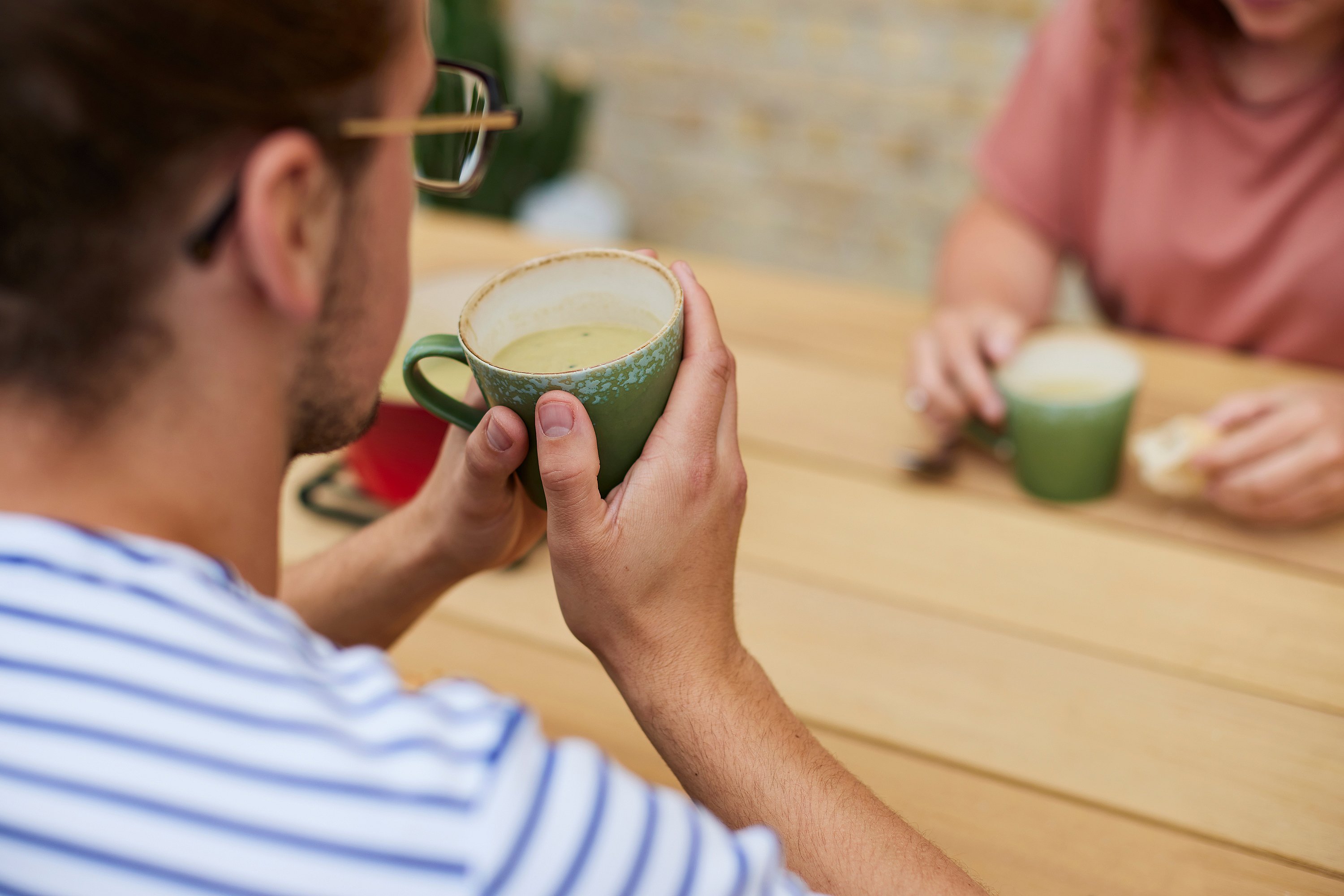 Nahaufnahme von hinten: Ein Mann hält einen grünen Becher Suppe über den Holztisch eines anderen Restaurants, dazwischen Brot und Geschirr.