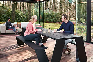 Twee collega's zitten tegenover elkaar aan een zwarte buitentafel in een paviljoen met glazen wanden, terwijl een derde persoon op een laptop werkt vanaf een beige bank op de achtergrond, met daarachter zichtbaar bos.