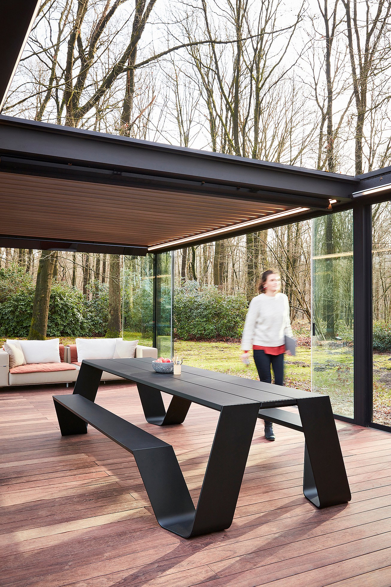 Schwarzer Außentisch mit passender Bank auf einer Holzterrasse im Glaspavillon, mit einem beigen Sofa im Hintergrund und einer Frau, die an der offenen Glaswand vorbeigeht.