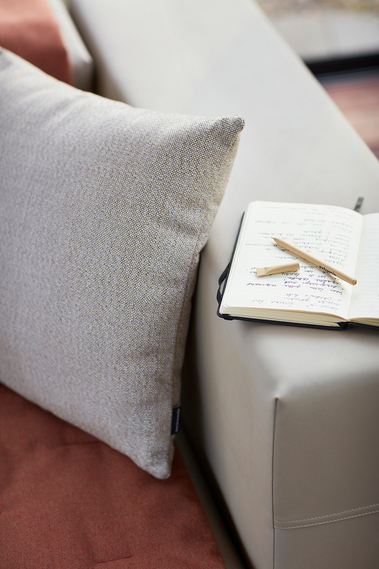 Nahaufnahme eines strukturierten hellgrauen Kissens neben einem offenen Notizbuch mit handschriftlichen Notizen und zwei Bleistiften, die auf dem Arm eines beigen Walrosssofas ruhen.
