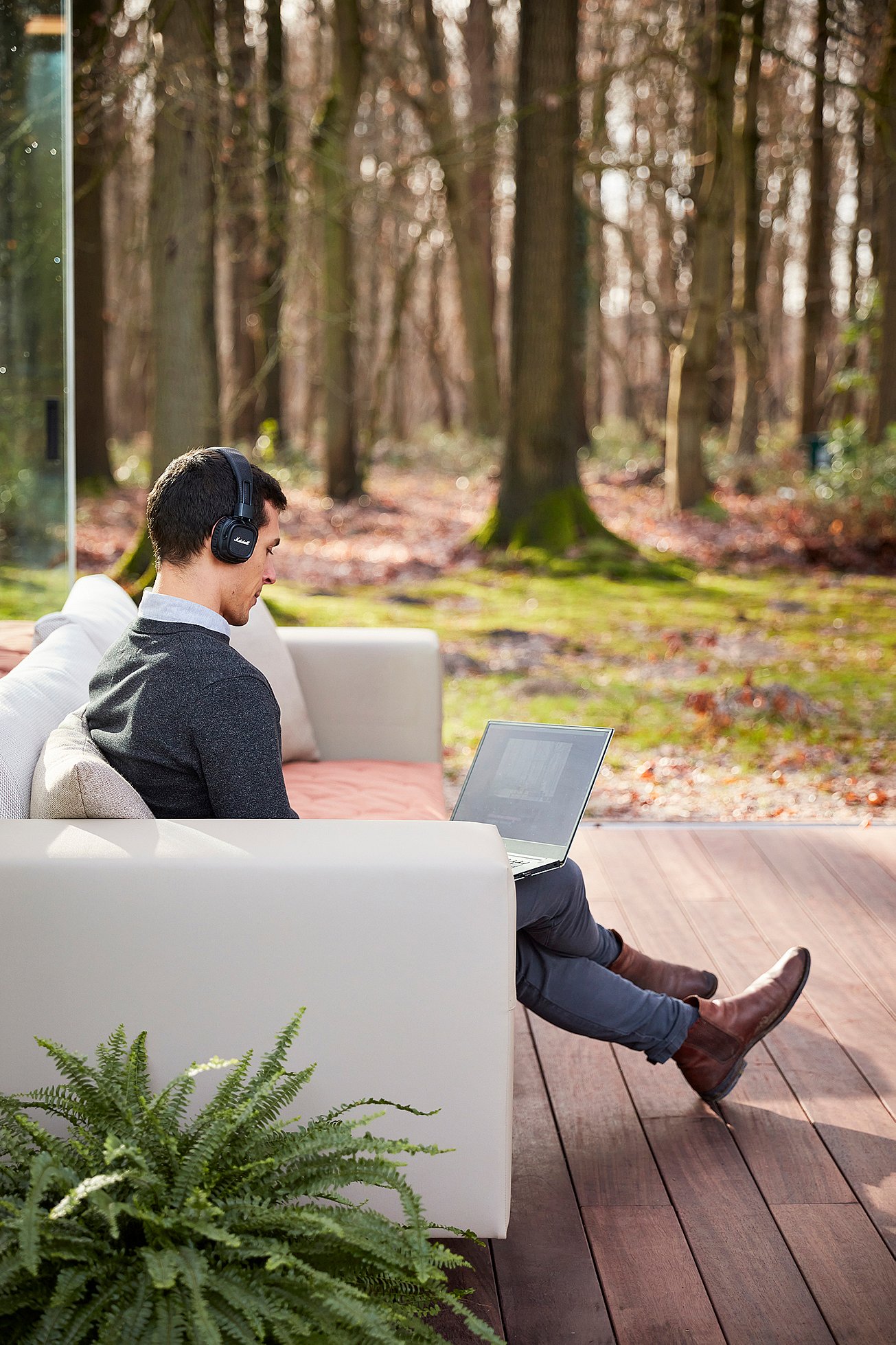   Ein Mann mit Kopfhörern arbeitet an einem Laptop, während er auf einem weißen Walrus-Sofa auf einer Holzterrasse mit Blick auf einen Wald sitzt.