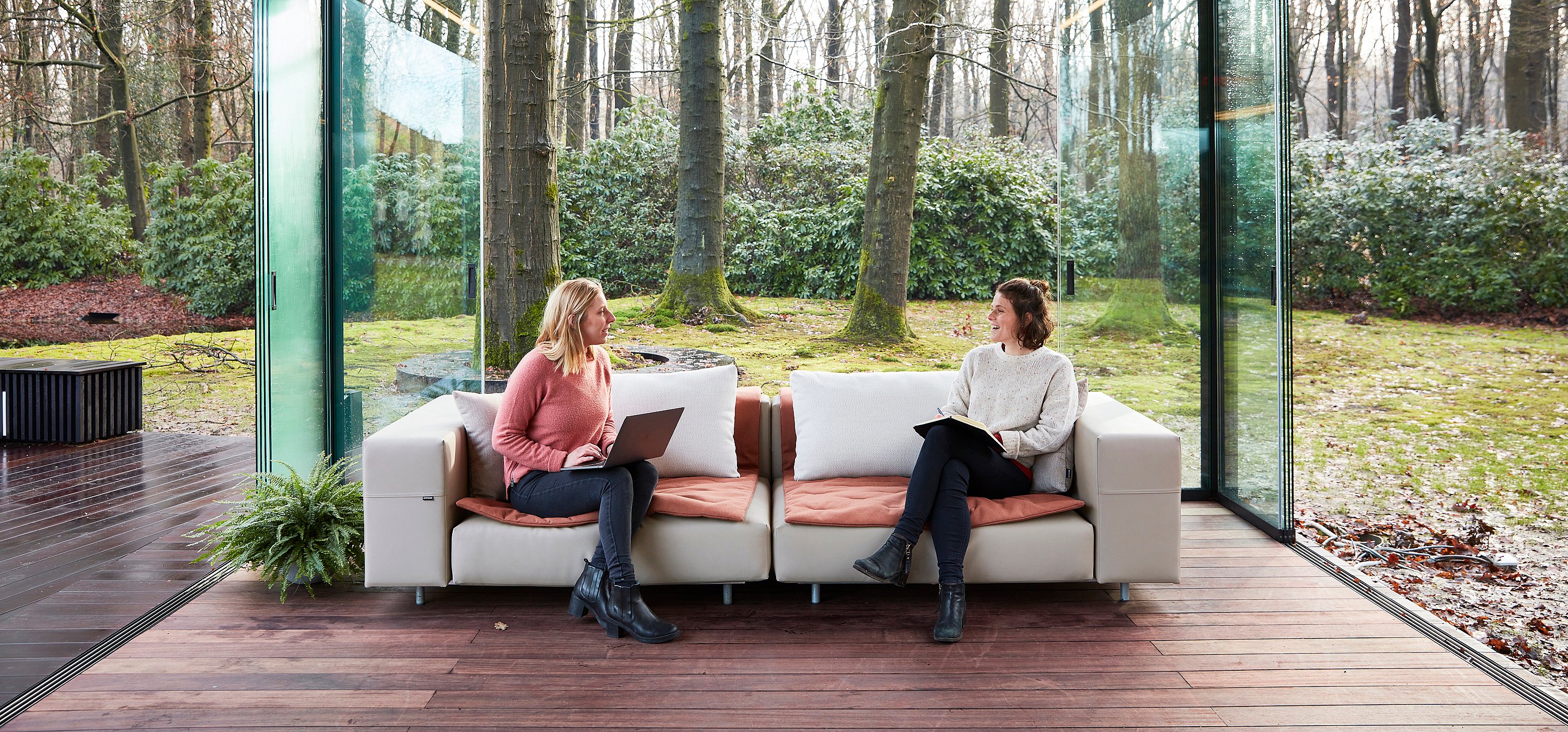 Zwei Frauen sitzen auf einem beigen modularen Walrosssofa mit Sitzkissen aus Terrakotta und weißen Rückenkissen in einem offenen Glaspavillon und arbeiten und sprechen mit Blick auf den Wald dahinter.