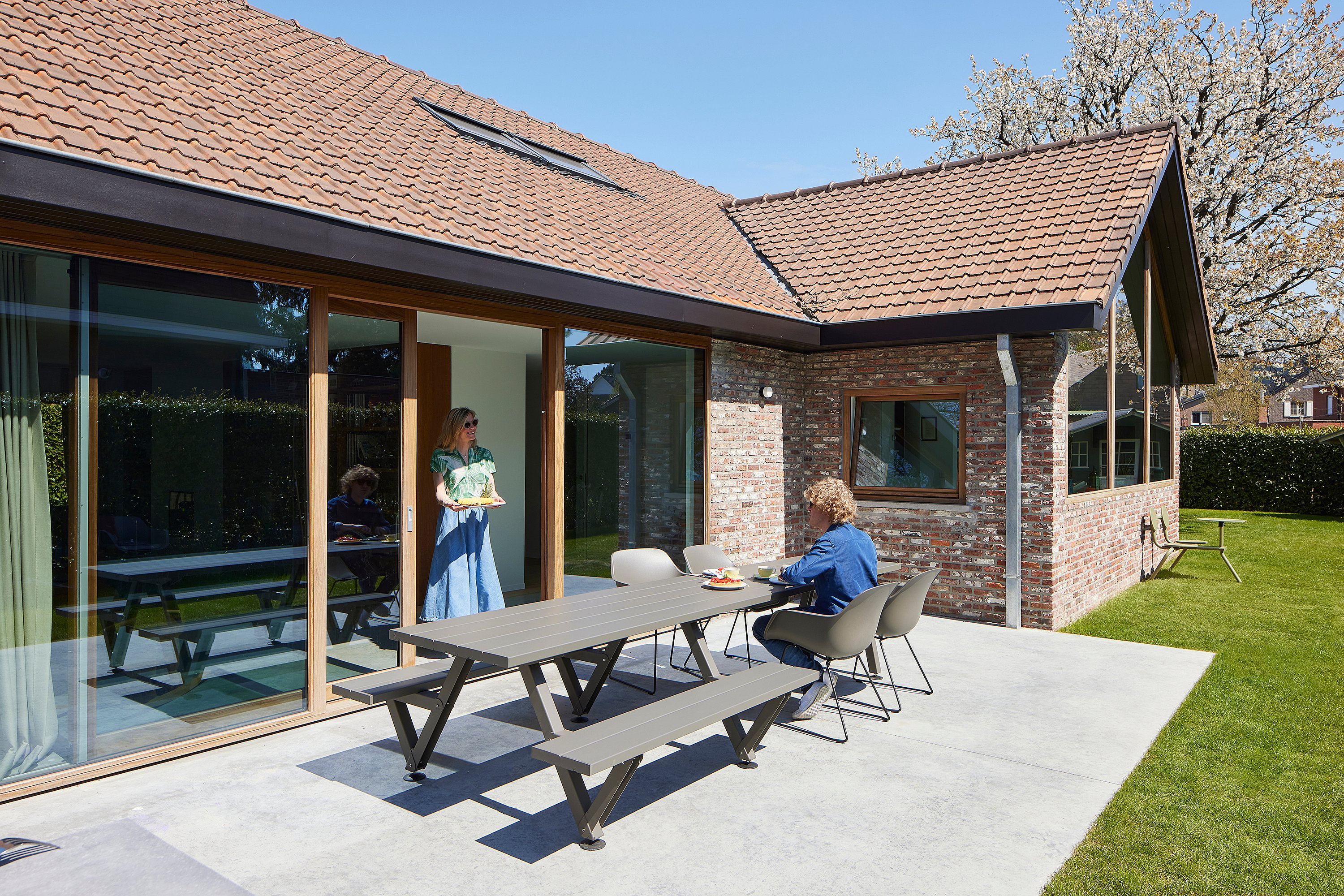 A sunlit brick house opens onto a patio with a long grey Marina outdoor table and Captain's Chairs, where one person sits and another stands in the doorway holding a tray.