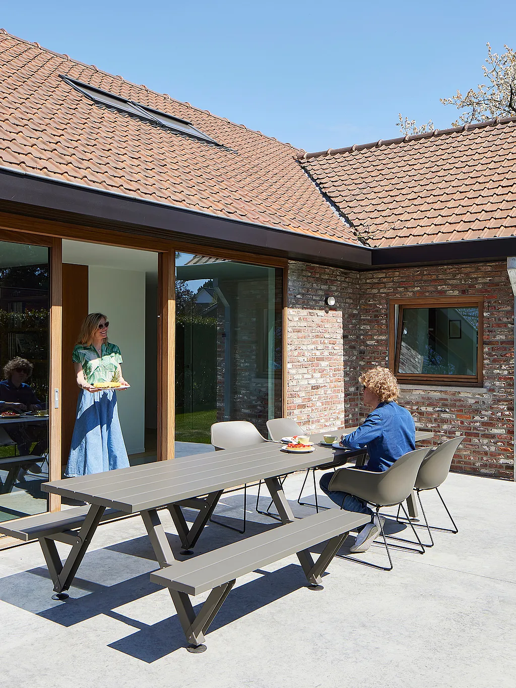 A sunlit brick house opens onto a patio with a long grey Marina outdoor table and Captain's Chairs, where one person sits and another stands in the doorway holding a tray.