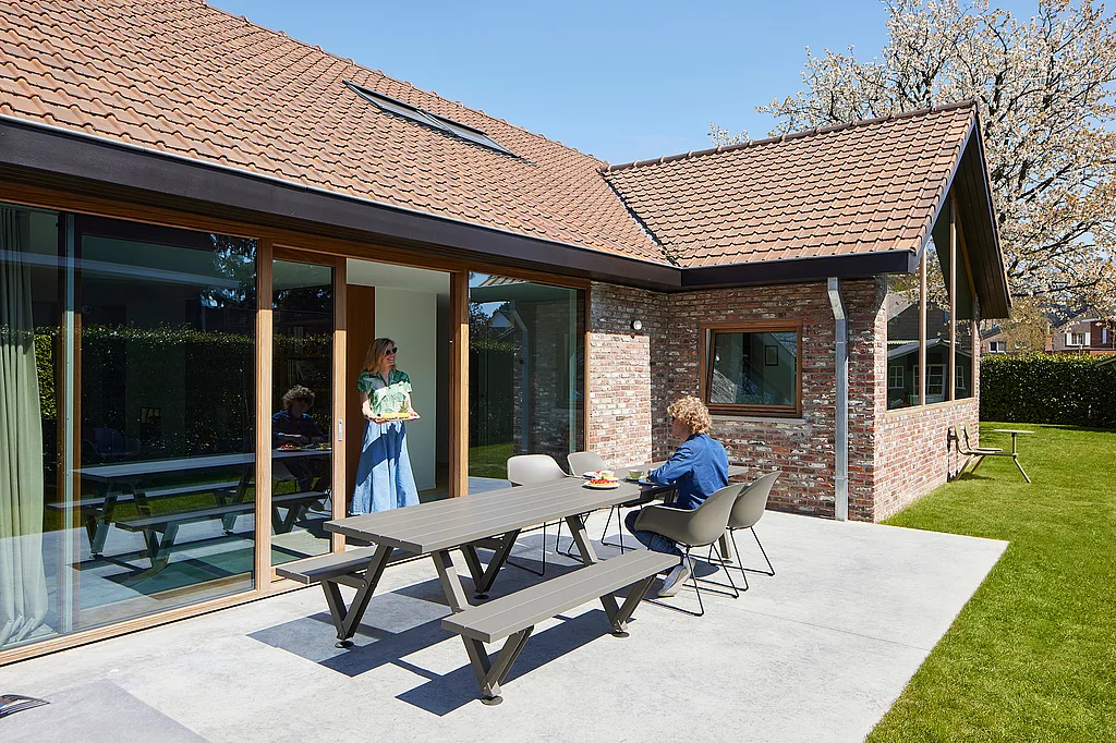 Une maison en briques baignée de soleil s'ouvre sur une terrasse où sont disposées une longue table d'extérieur grise de marque Marina et des chaises de style capitaine. Une personne est assise tandis qu'une autre, debout dans l'embrasure de la porte, tient un plateau.