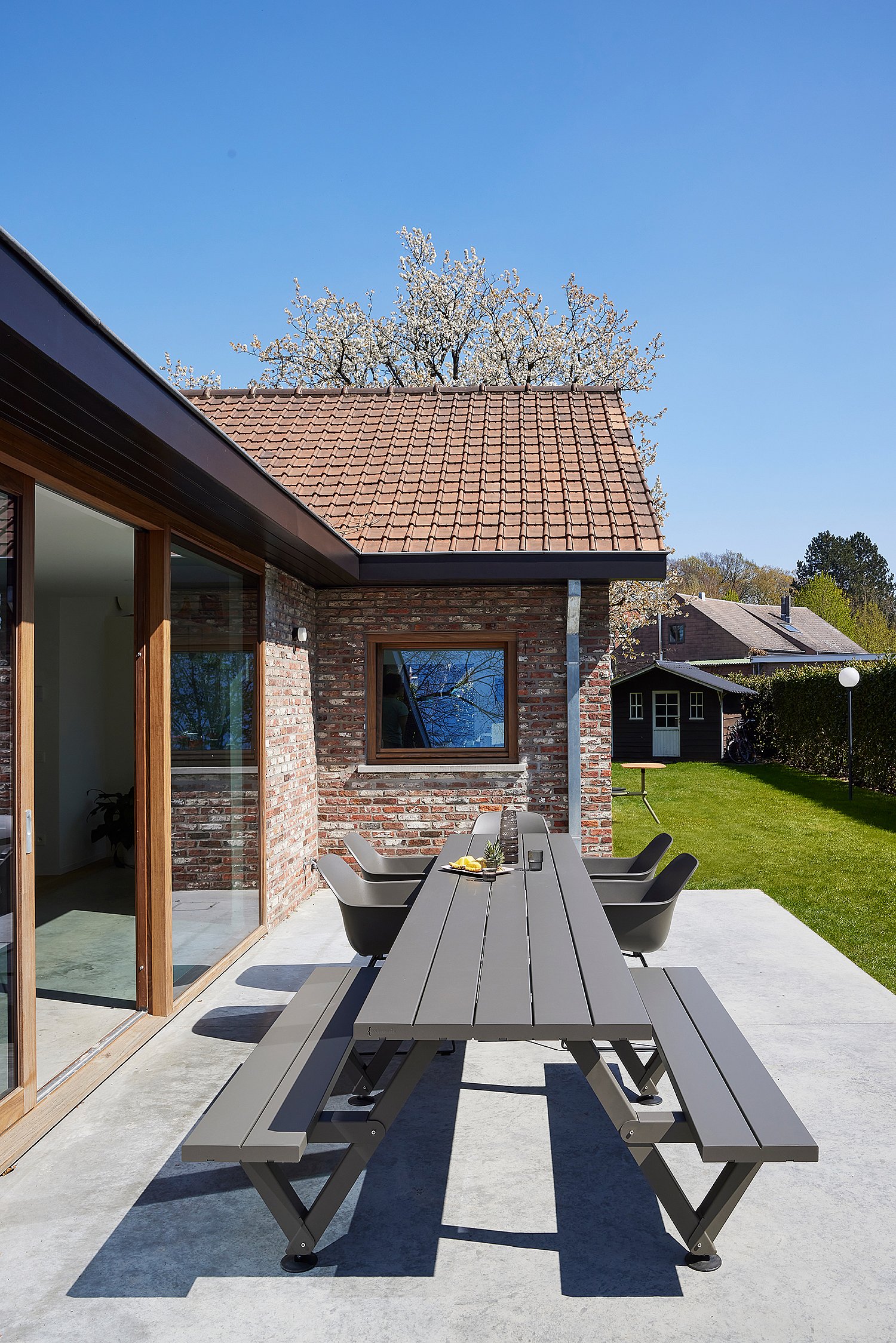 A long grey outdoor Marina table with benches and Captain's chairs stands on a patio beside a brick house and green lawn under a clear blue sky.