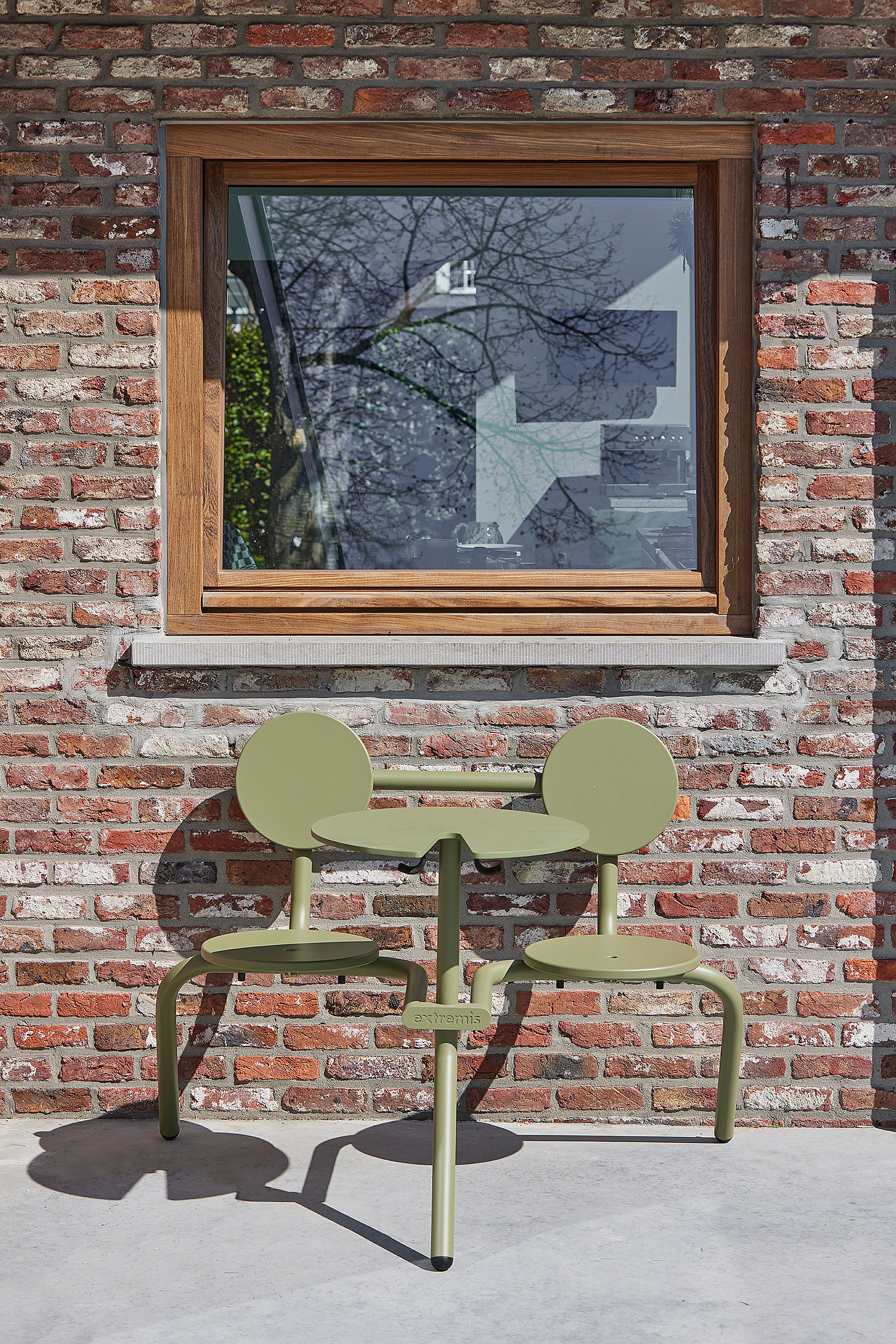 A compact green outdoor table with two attached seats stands against a brick wall below a wooden-framed window