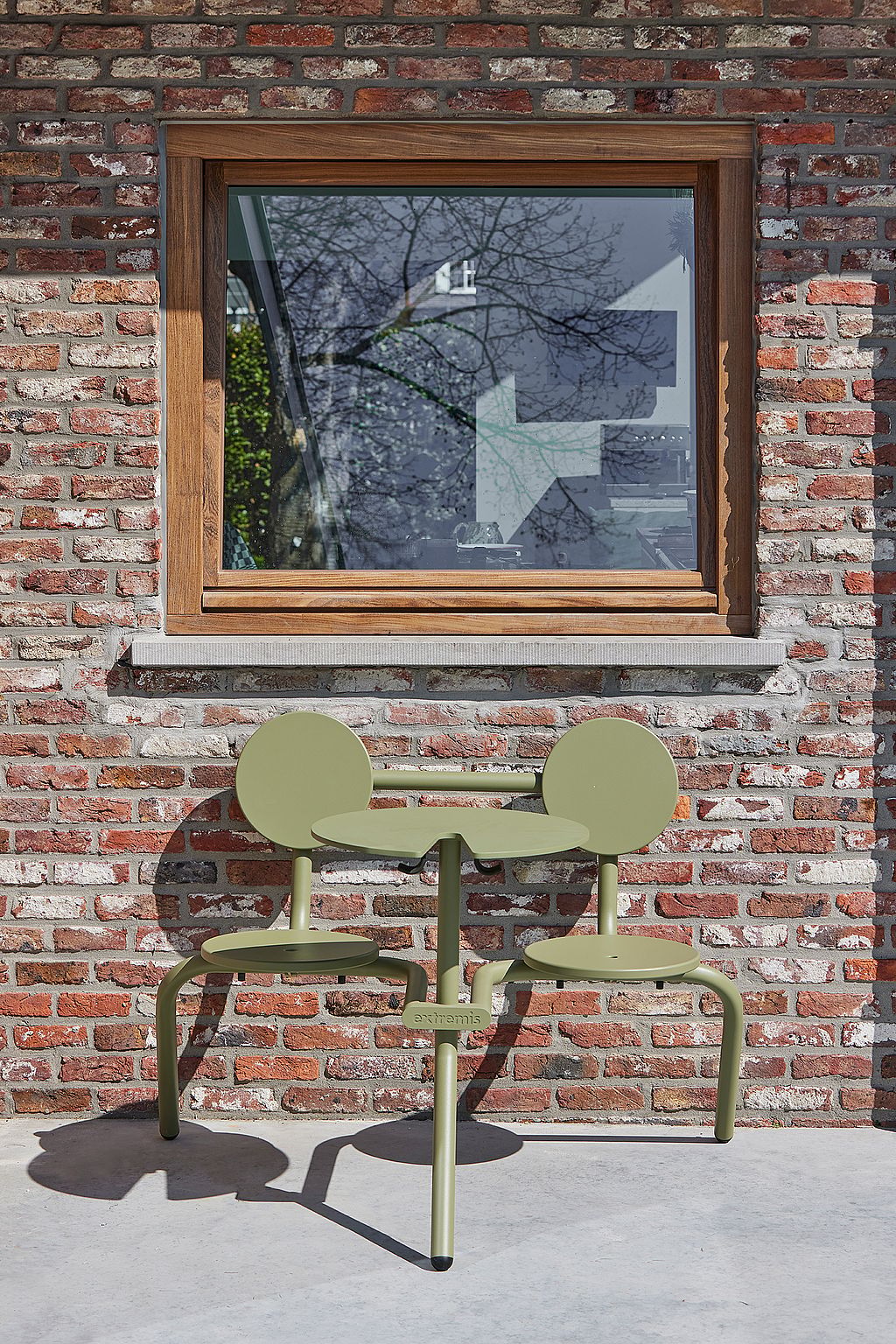 A compact green outdoor table with two attached seats stands against a brick wall below a wooden-framed window