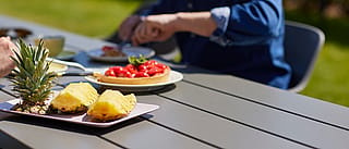 Close-up of fruit and dessert on a grey outdoor table, with two people blurred in the background enjoying a meal outside.