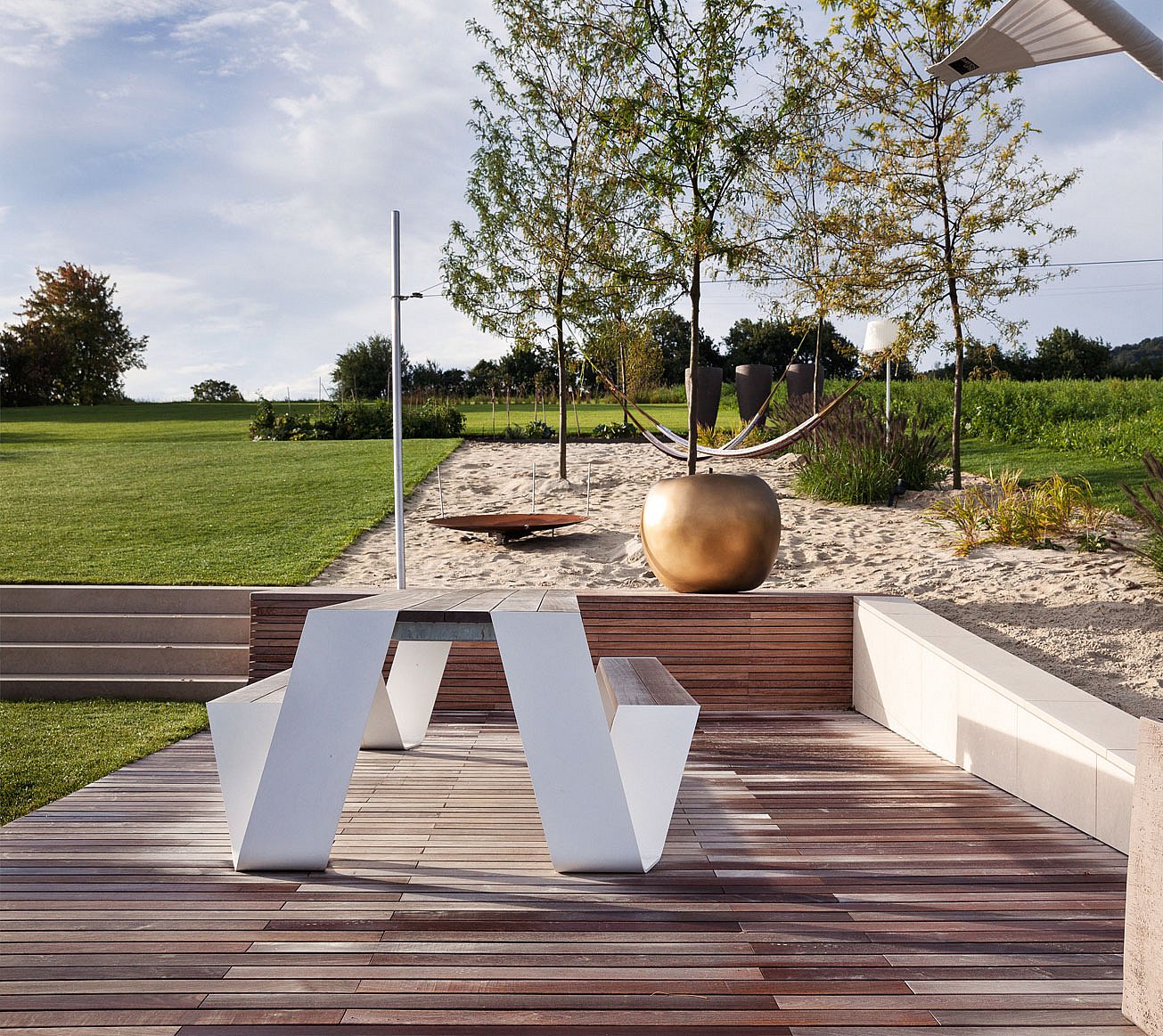 Witte picknicktafel op een houten terras voor een zandtuin met een hangmat tussen kleine bomen en een bronzen planter in het midden.