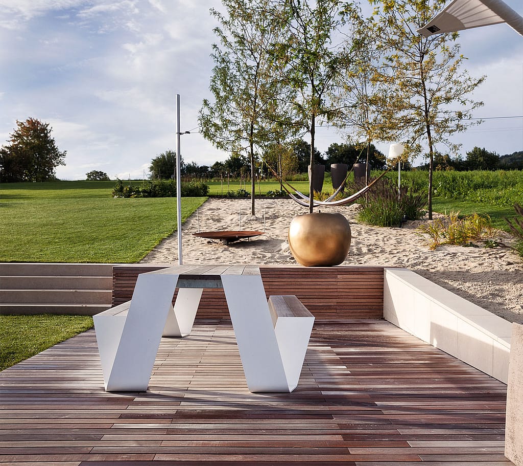 Witte picknicktafel op een houten terras voor een zandtuin met een hangmat tussen kleine bomen en een bronzen planter in het midden.