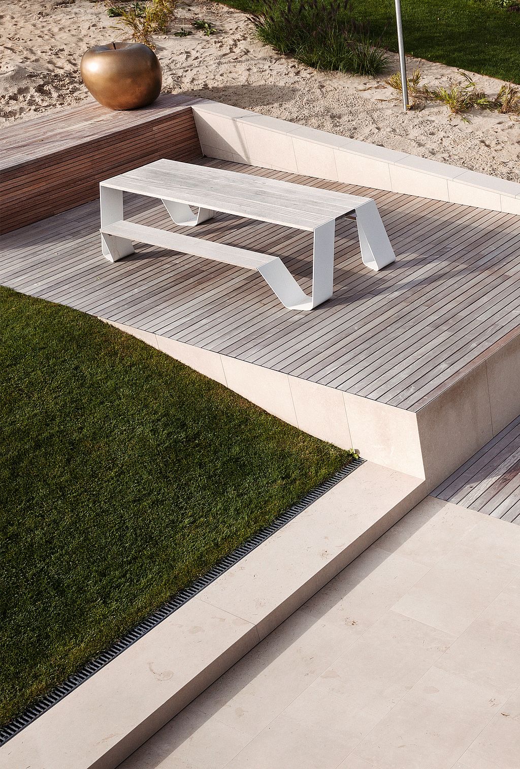 Witte hopper picknicktafel met aangebouwde bank op een houten terras naast een zandtuin, met een grote bronzen planter aan de achterkant.