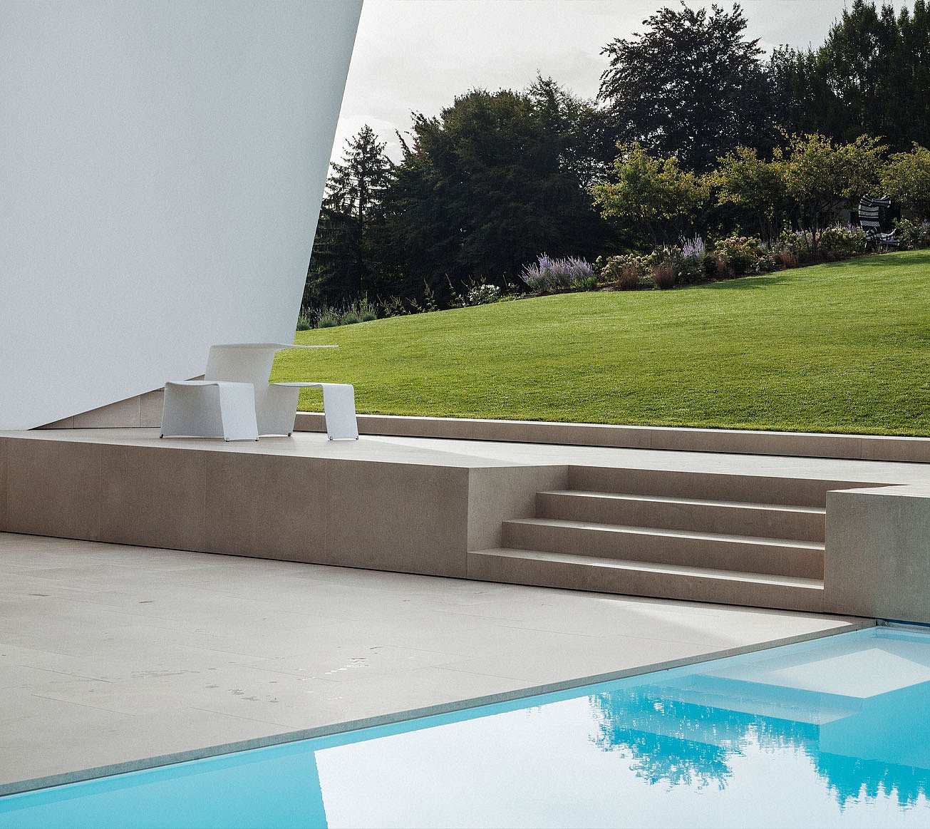 Minimalistische weiße Terrasse neben einem stillen Swimmingpool, mit hellen Steinstufen, die zu einer abfallenden Rasenfläche führen, und einem weißen Picknick im Freien unter dem scharf abgewinkelten Überhang des Hauses.