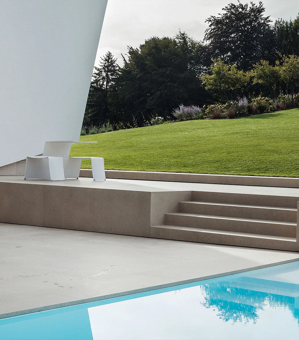 Terrasse blanche minimaliste à côté d’une piscine immobile, avec des marches en pierre pâle menant à une pelouse en pente et un pique-nique extérieur blanc sous le surplomb fortement incliné de la maison.