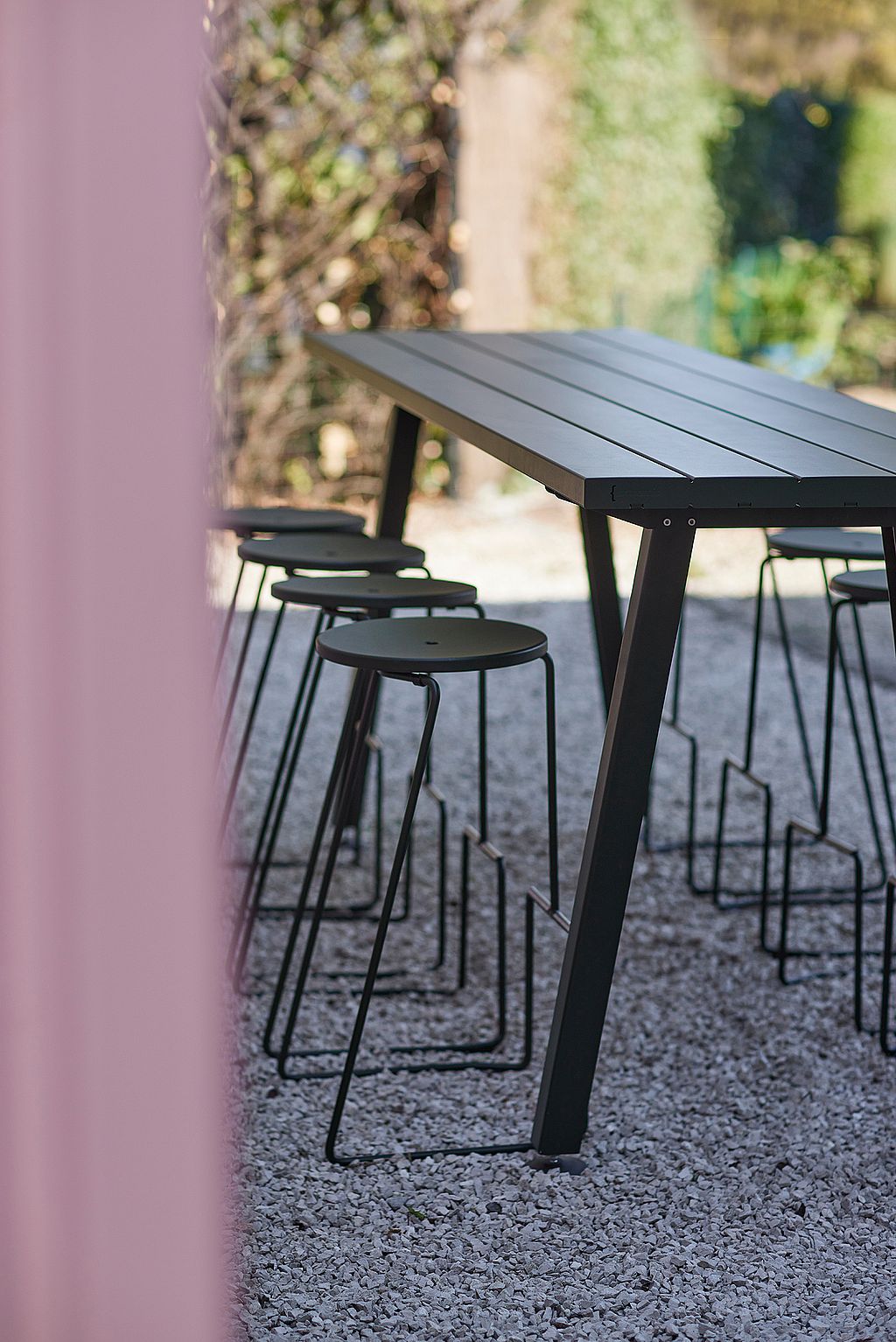 Zijaanzicht van een op maat gemaakte Marina-tafel met een rij Tiki-barkrukken op grind naast een roze muur, gefotografeerd in zacht buitenlicht.