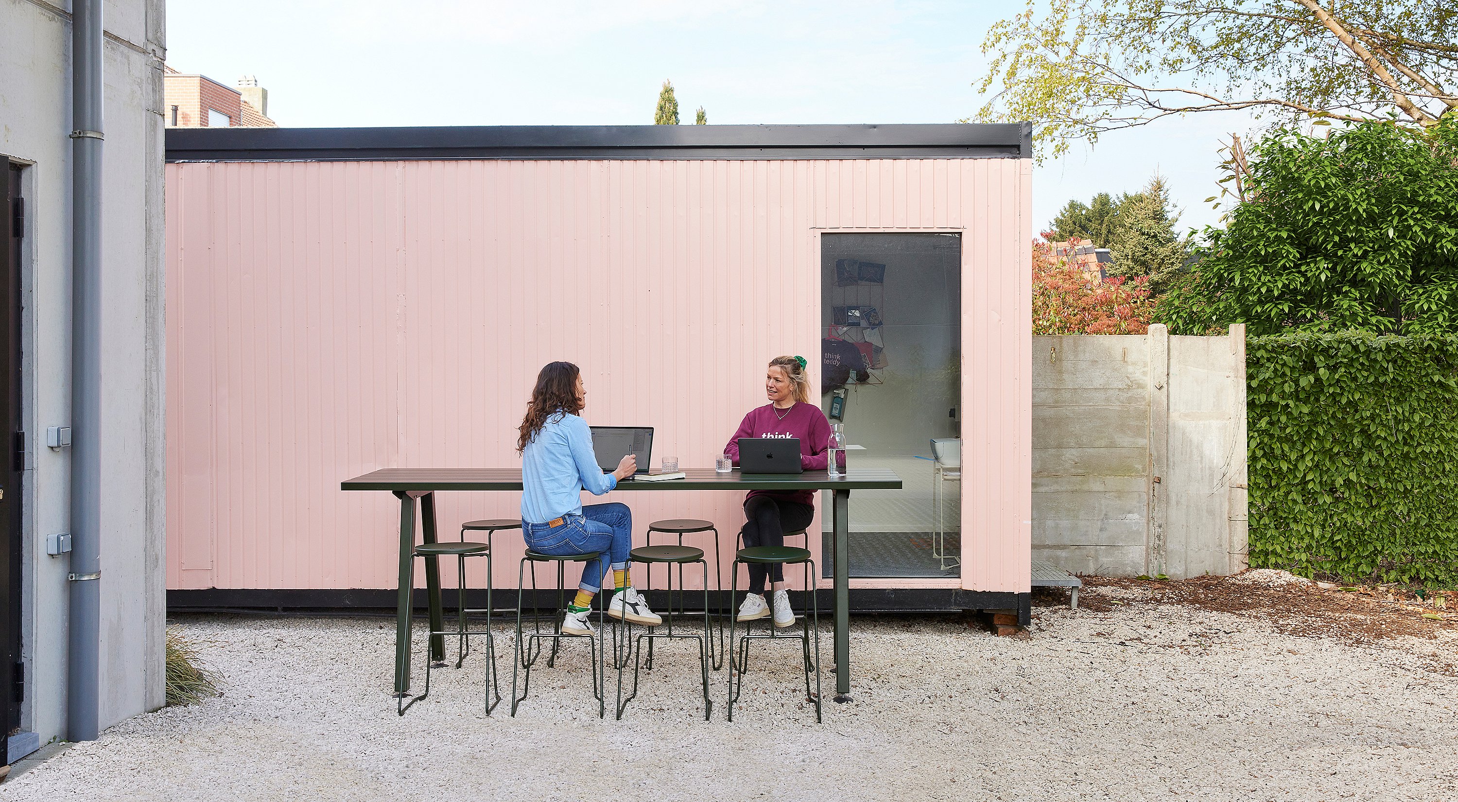 Twee vrouwen werken buiten aan een op maat gemaakte Marina hoge tafel met Tiki barkrukken voor een roze containerkantoor.