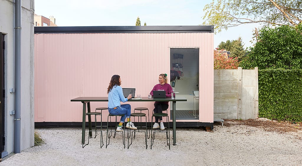 Twee vrouwen werken buiten aan een op maat gemaakte Marina hoge tafel met Tiki barkrukken voor een roze containerkantoor.