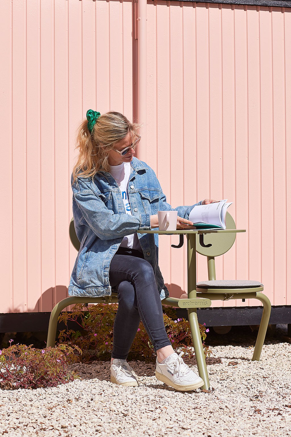 Een vrouw leest en schrijft aan een groene Bistroo tafel met vaste stoelen, geplaatst op wit grind voor een roze buitenmuur.