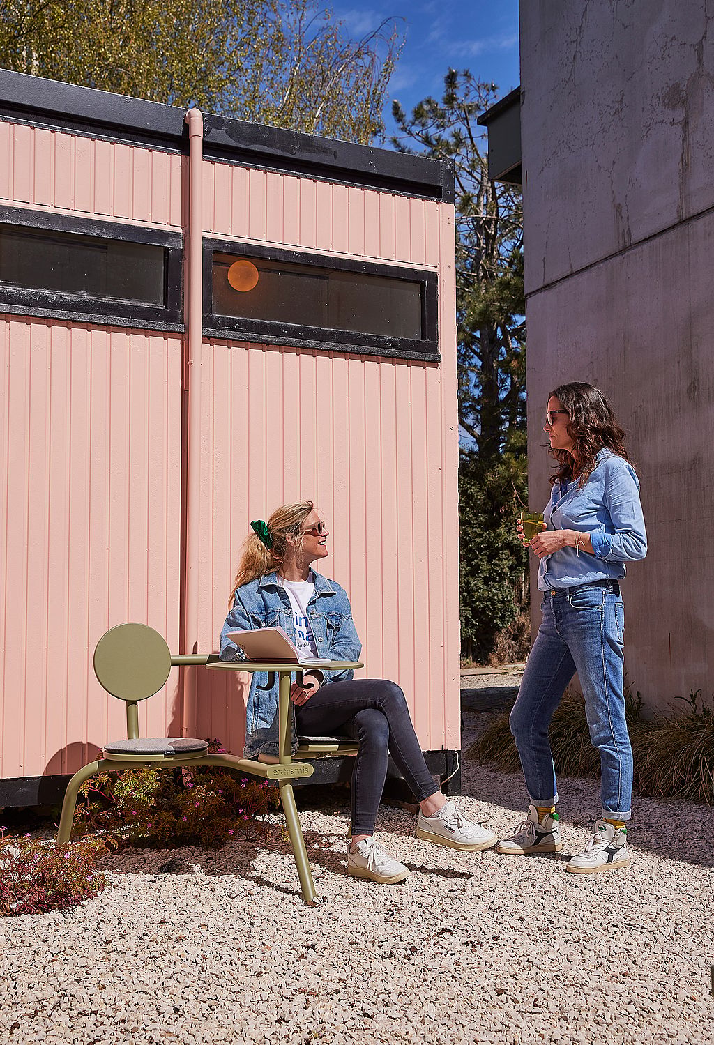 Twee vrouwen kletsen naast een groene Bistroo tafel met vaste stoelen in een zonnige buitenwerkplek naast een roze containergebouw.