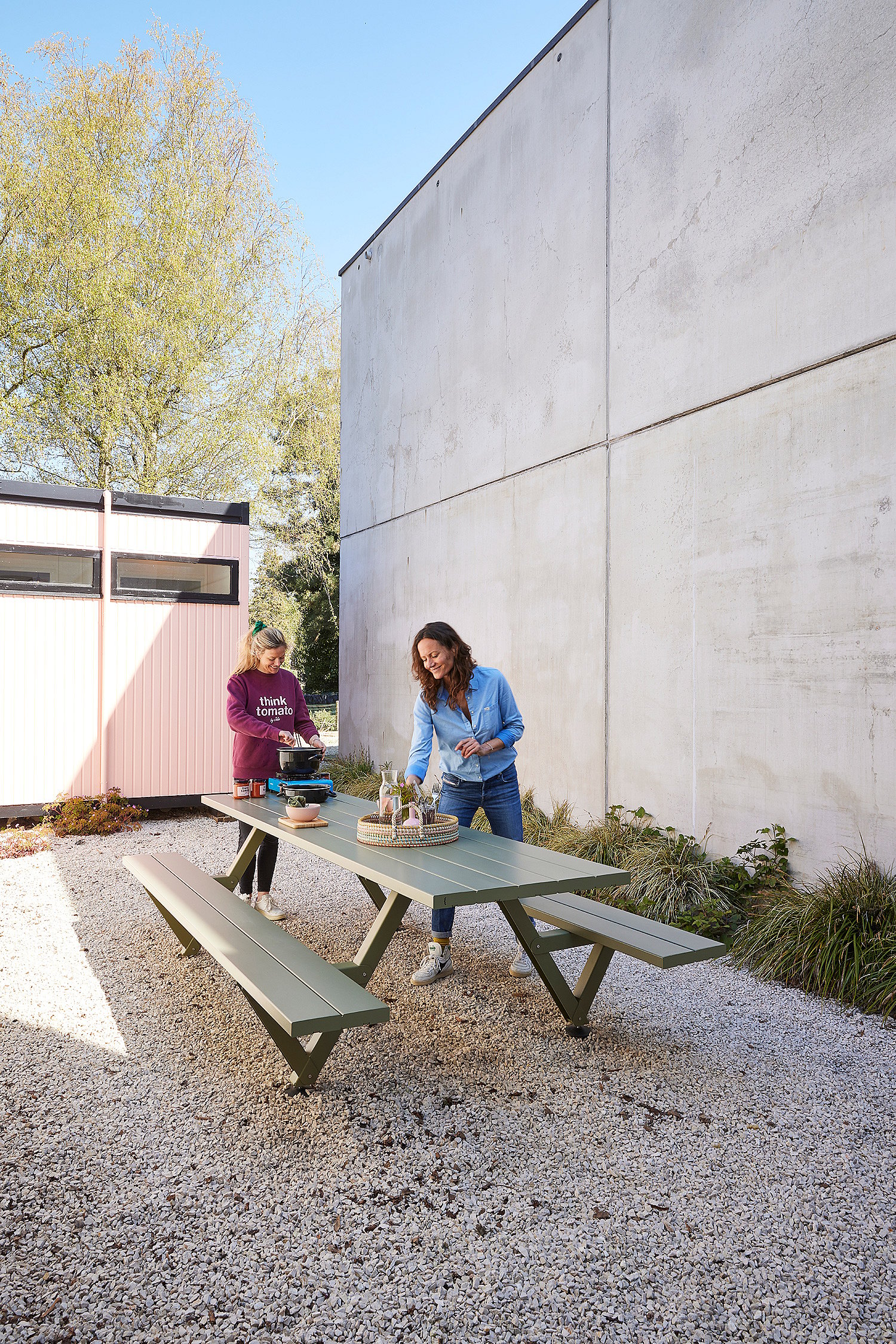 Twee vrouwen bereiden eten en zetten drankjes klaar op een Reed groene Marina picknicktafel in een minimalistische buitenpatio.
