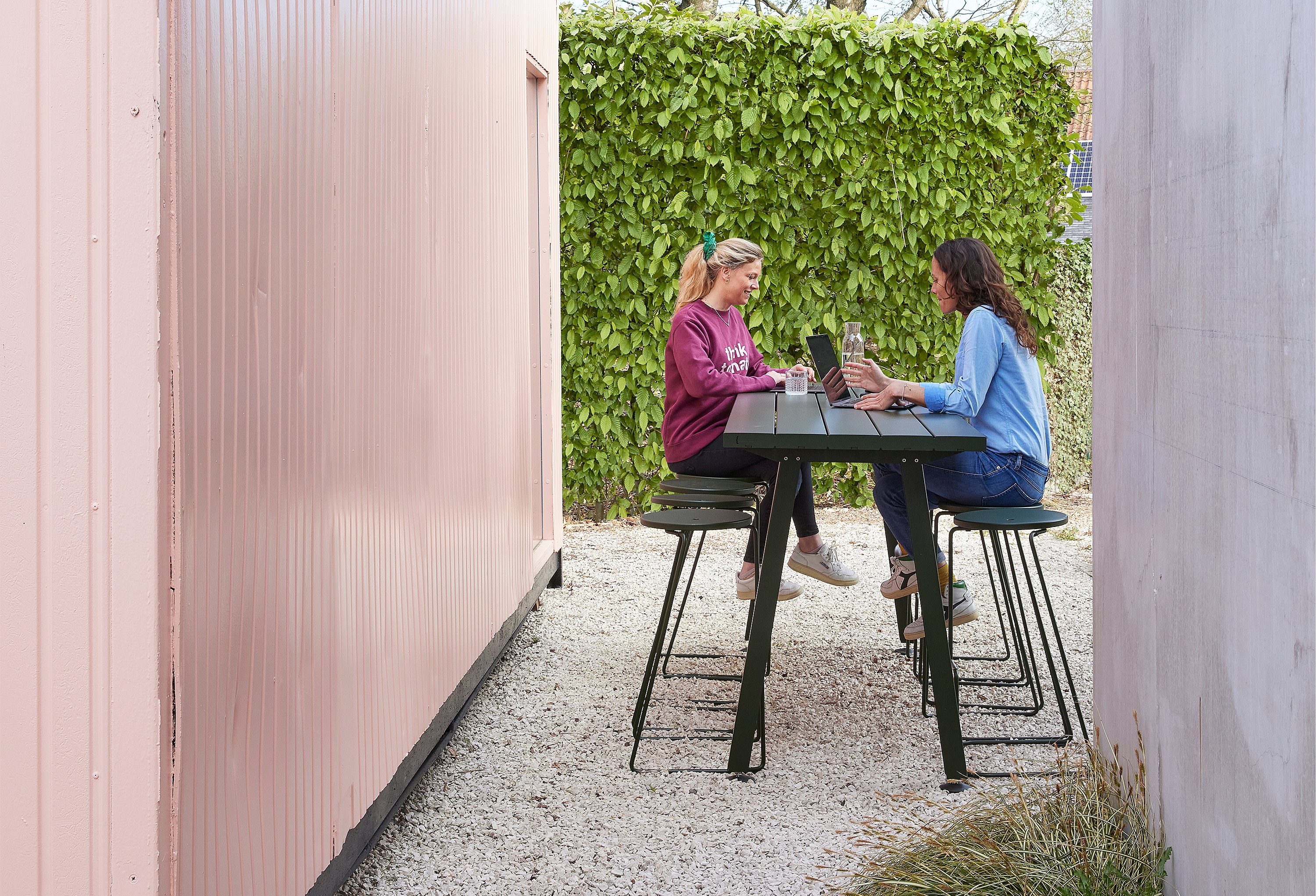 Twee vrouwen zitten tegenover elkaar aan een op maat gemaakte Marina-tafel met Tiki-barkrukken, werkend op hun laptops in een grindtuin tussen een roze containerwand en een hoge groene heg.