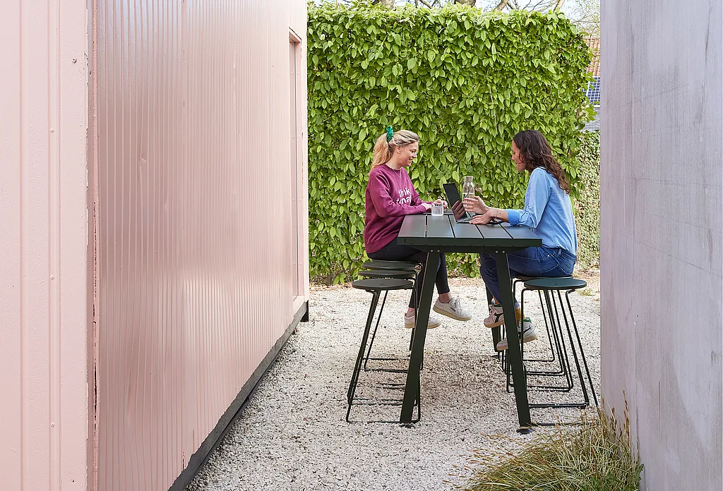 Two women sit facing each other at a custom-coloured Marina high table with Tiki bar stools, working on laptops in a gravel courtyard between a pink container wall and a tall green hedge.