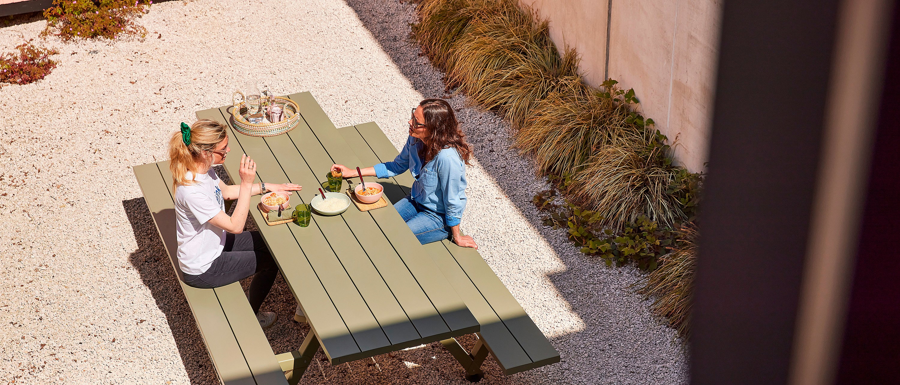   Zwei Frauen sitzen an einem grünen Picknicktisch in einem Kieshof, essen zu Mittag und unterhalten sich im hellen Sonnenlicht.