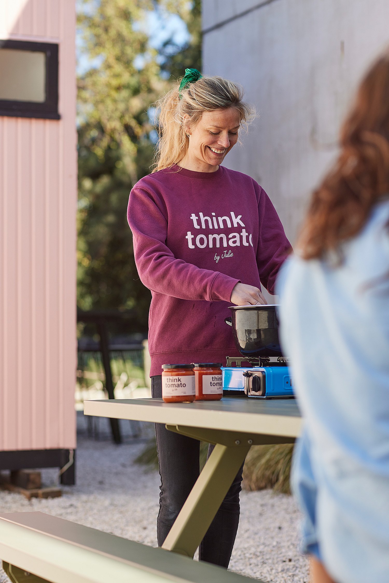   Een vrouw kookt buiten aan een groene picknicktafel in Reed Green Marina, met potten Think Tomato saus en een draagbaar kooktoestel op tafel.