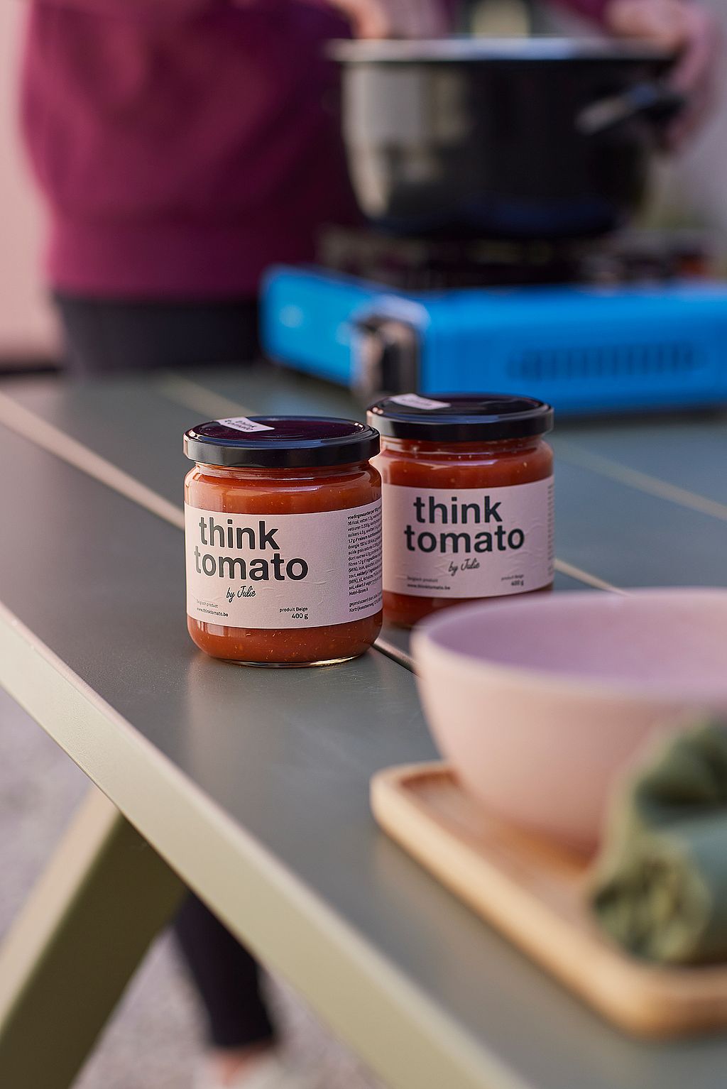 Close-up van twee potten Think Tomato saus op een groene picknicktafel in Reed Green Marina, met een kom en een draagbaar kooktoestel wazig op de achtergrond.