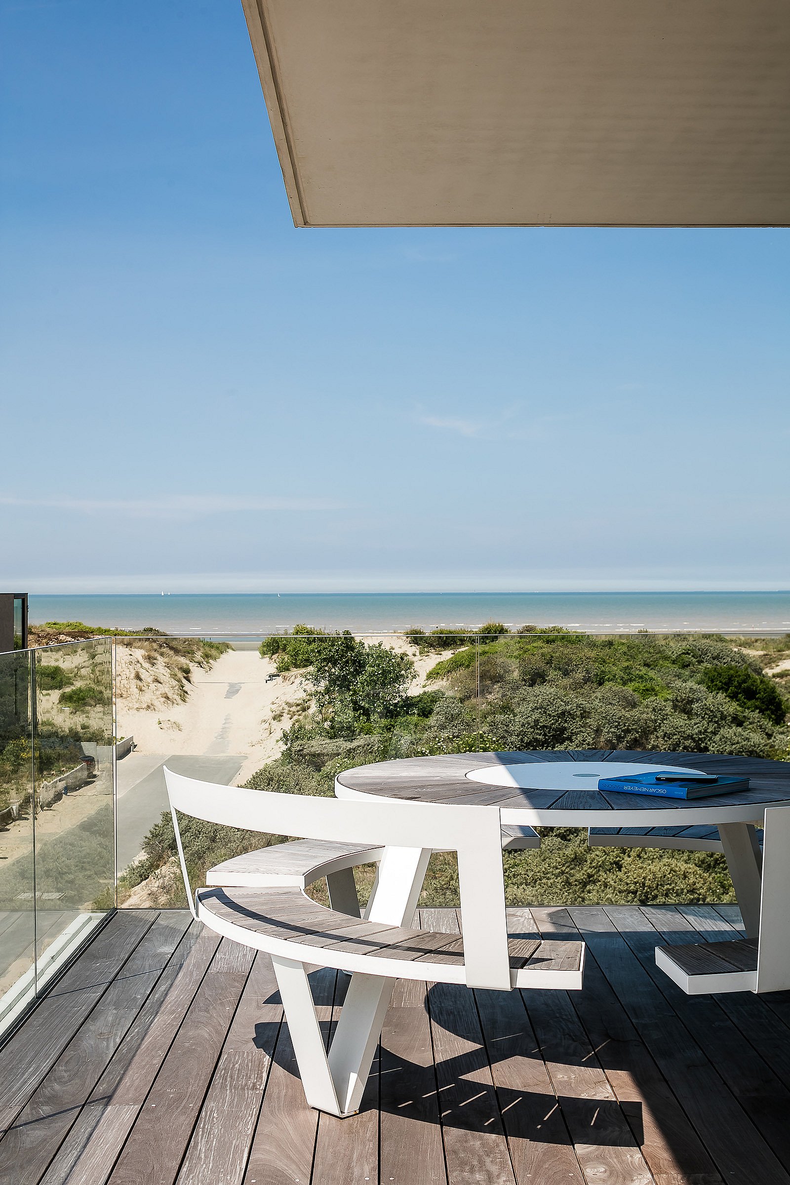 Close view of a white round outdoor pantagruel picnic table with curved bench seats on a wooden terrace, overlooking dune vegetation, a sandy path to the beach, and the sea beyond.