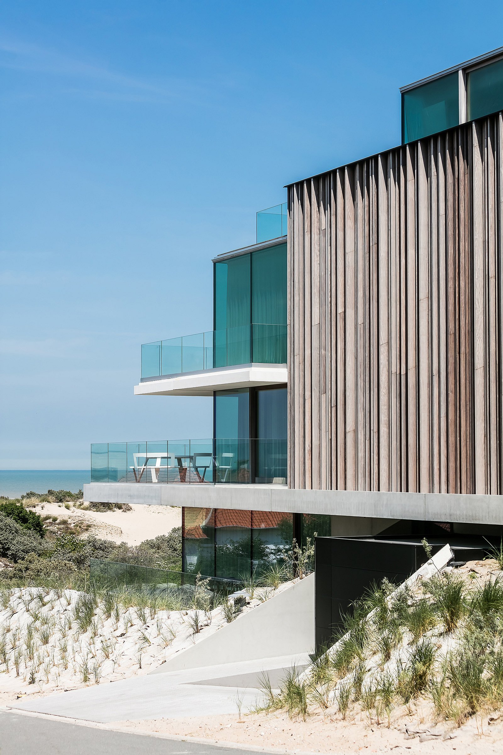 Modern coastal house set among sand dunes, with vertical timber cladding, glass balconies, and a white round pantagruel picnic table on a sea-facing terrace under a clear blue sky.