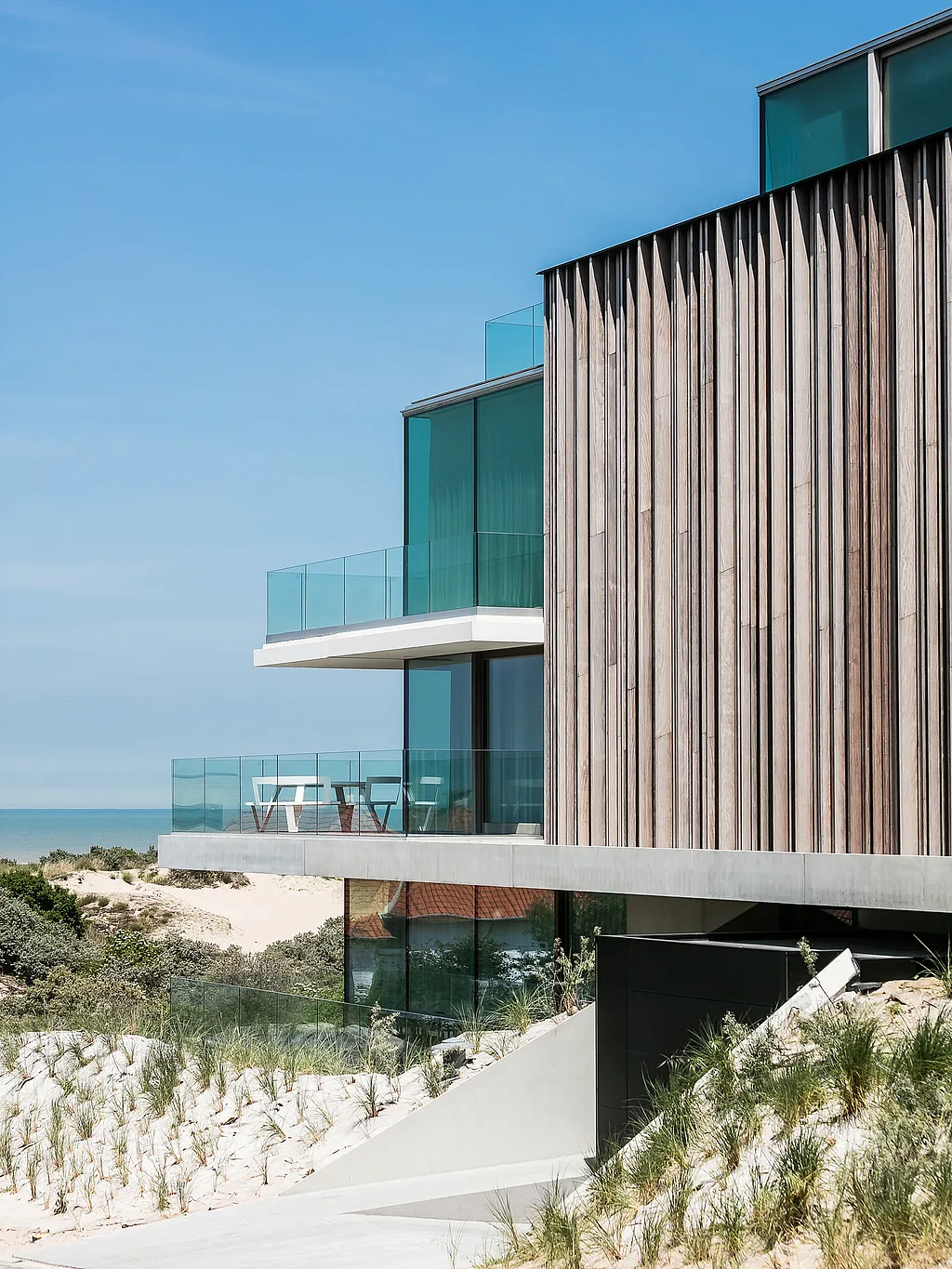 Modernes Küstenhaus inmitten von Sanddünen mit vertikaler Holzverkleidung, Glasbalkonen und einem weißen, runden Picknicktisch aus Pantagruel auf einer Terrasse mit Meerblick unter klarem, blauem Himmel.