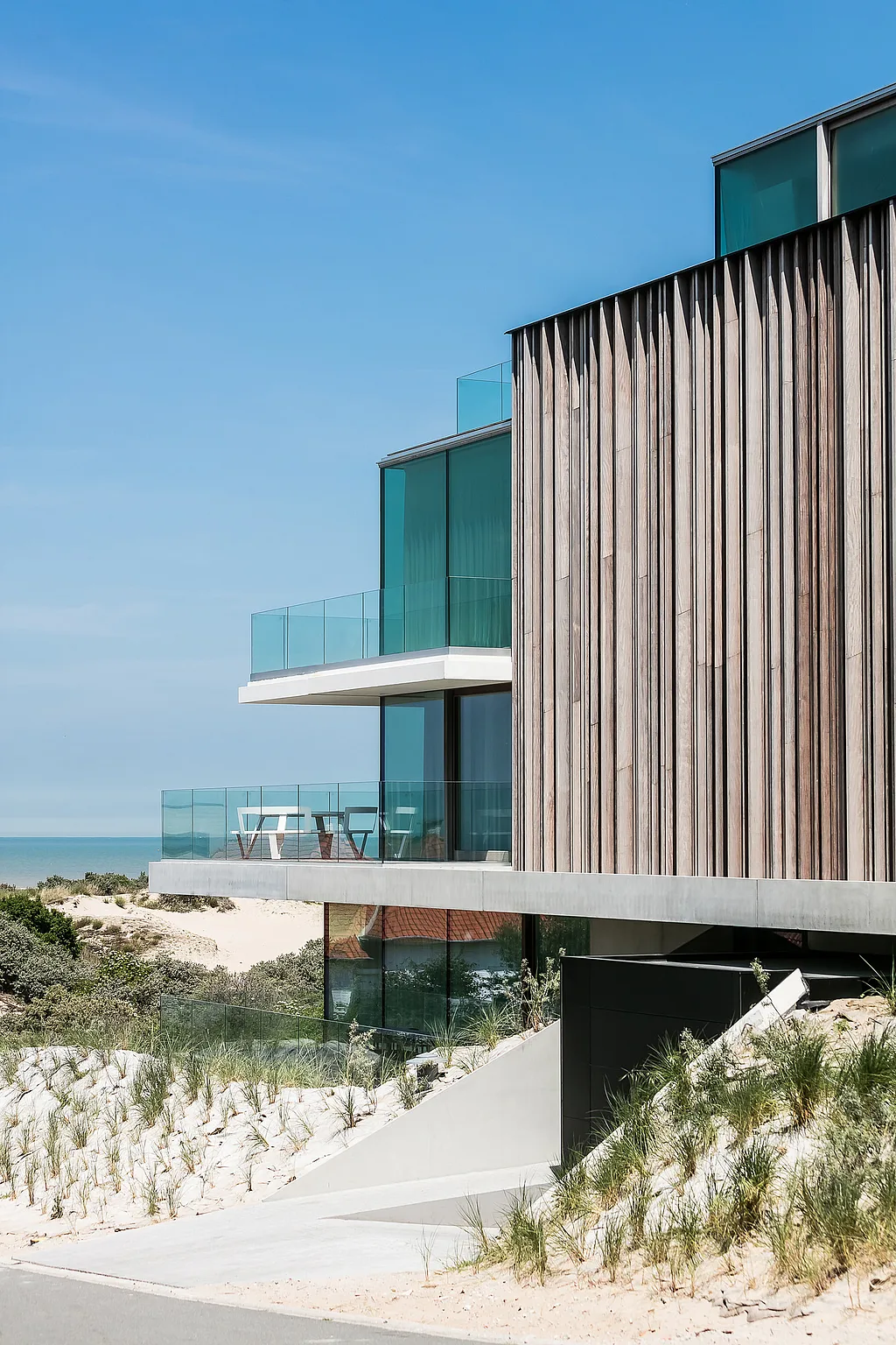 Modern coastal house set among sand dunes, with vertical timber cladding, glass balconies, and a white round pantagruel picnic table on a sea-facing terrace under a clear blue sky.