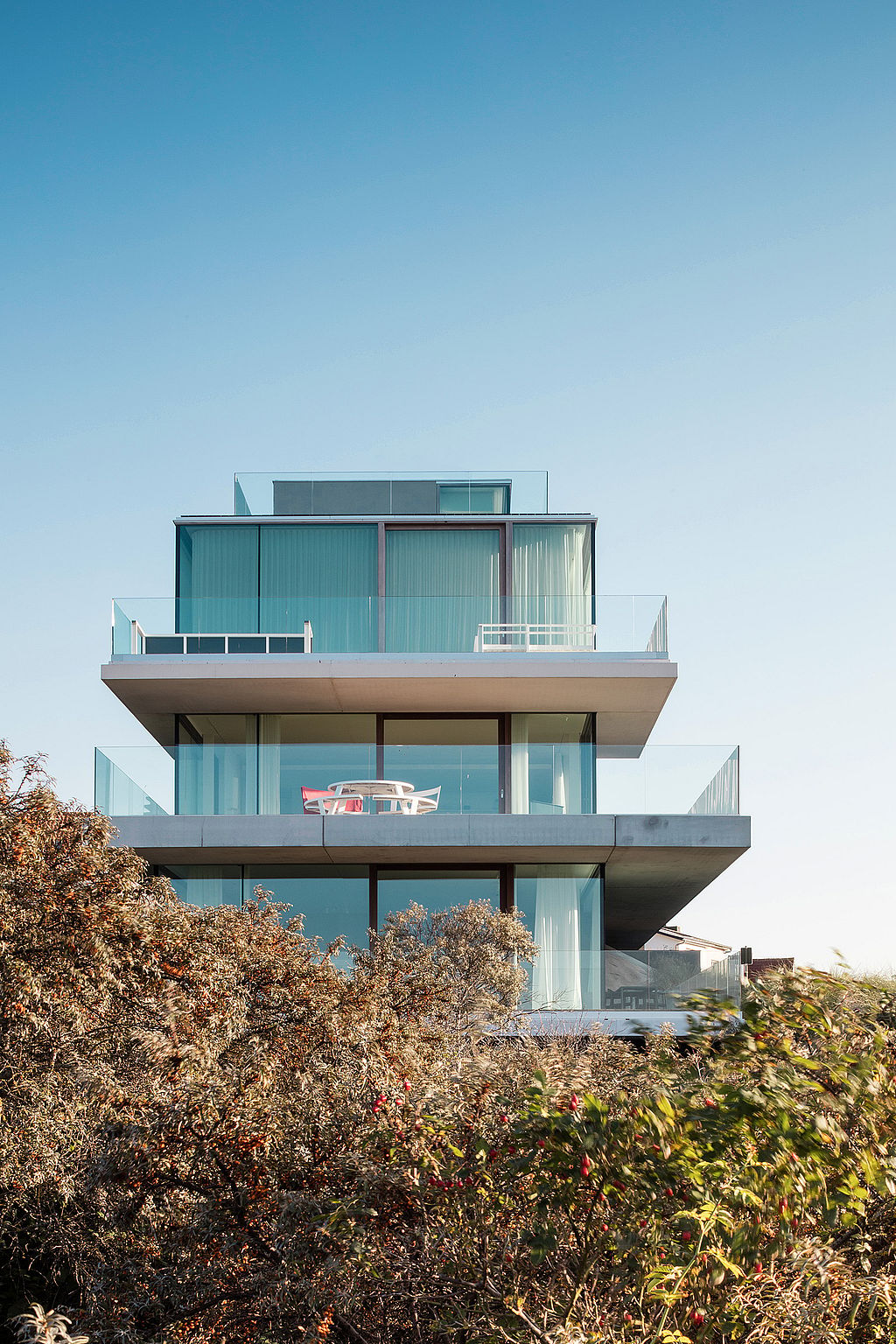 Front view of a modern multi-level beach house with wide glass façades and terraces, with a white round pantagruel picnic table visible on the middle balcony above the dunes.