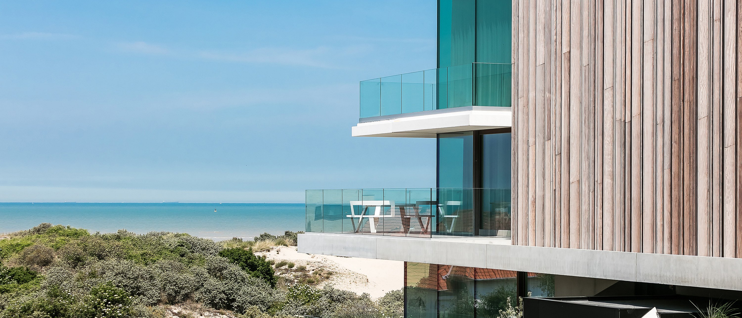 Side view of the timber-clad house with glass balconies above the dunes, with a white pantagruel round picnic table on the terrace and the North Sea stretching out behind it.