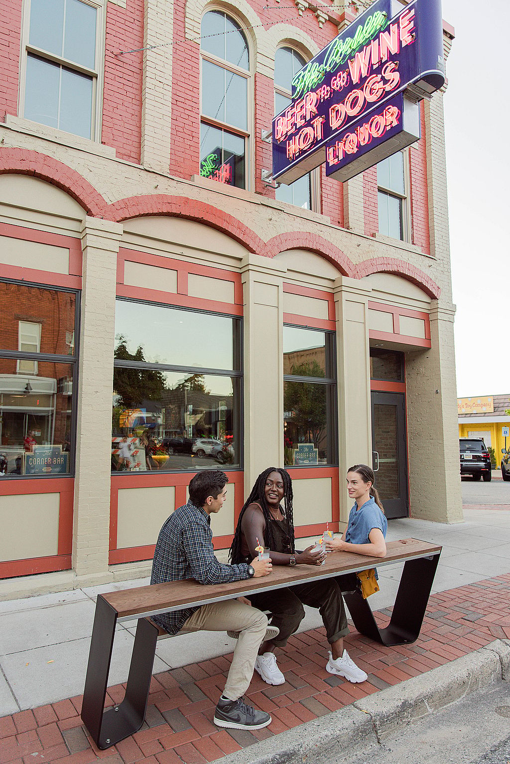 Trois amis sont assis sur un banc de hopper sur le trottoir sous une enseigne au néon vintage indiquant « Beer to go », « Wine », « Hot Dogs », « Liquor » dans le bâtiment d’angle derrière eux.