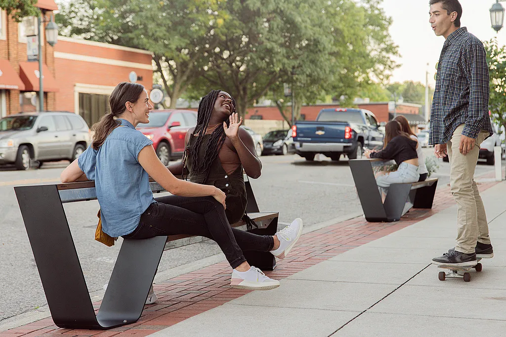Two women sit on a sidewalk hopper bench talking with a young man on a skateboard, while another pair sits on a second bench farther down the street beside parked cars and brick storefronts.