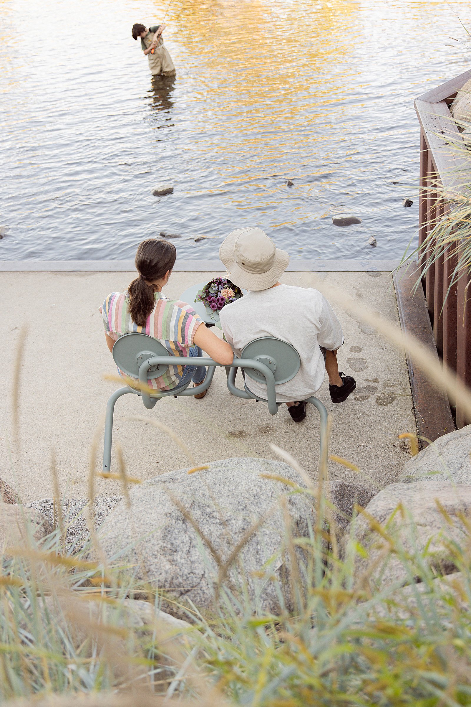 Rückansicht eines Paares, das auf einem Grünspan-Bistroo am Wasser sitzt und eine Person beobachtet, die im Fluss steht.