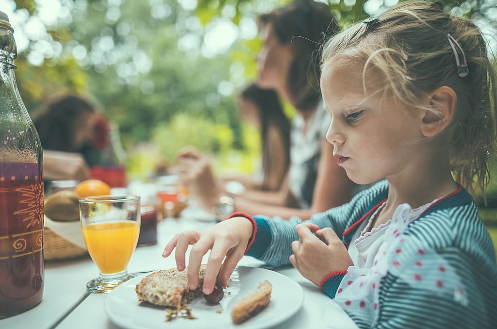 Close-up of a young girl eating breakfast at a Marina picnic table, with orange juice and family members softly out of focus.