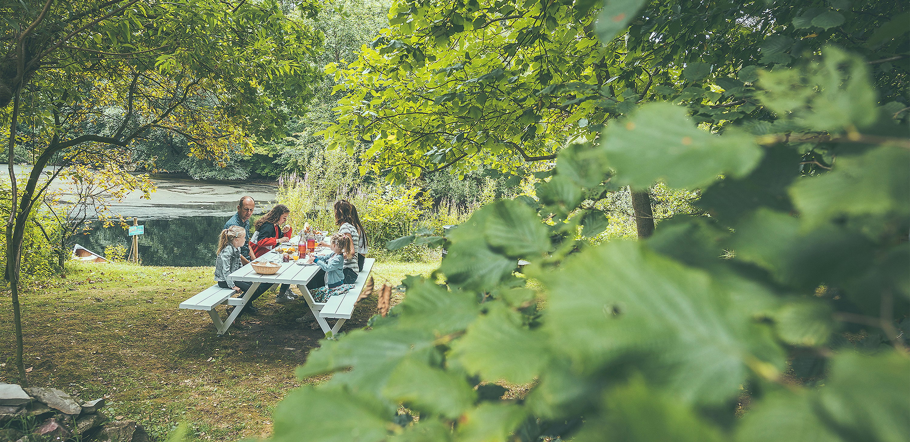 Seen through green leaves, a family shares breakfast at a Marina picnic table on the grassy lakeside.