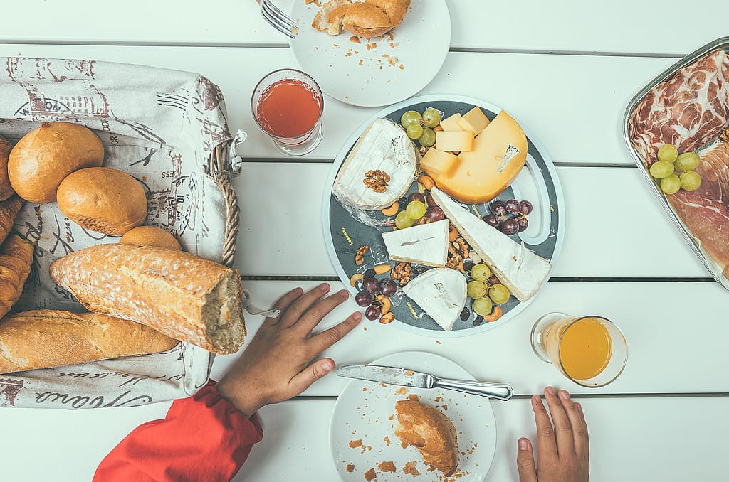 Overhead view of bread, cheese, charcuterie, grapes and juice spread across the pale surface of a Marina picnic table.