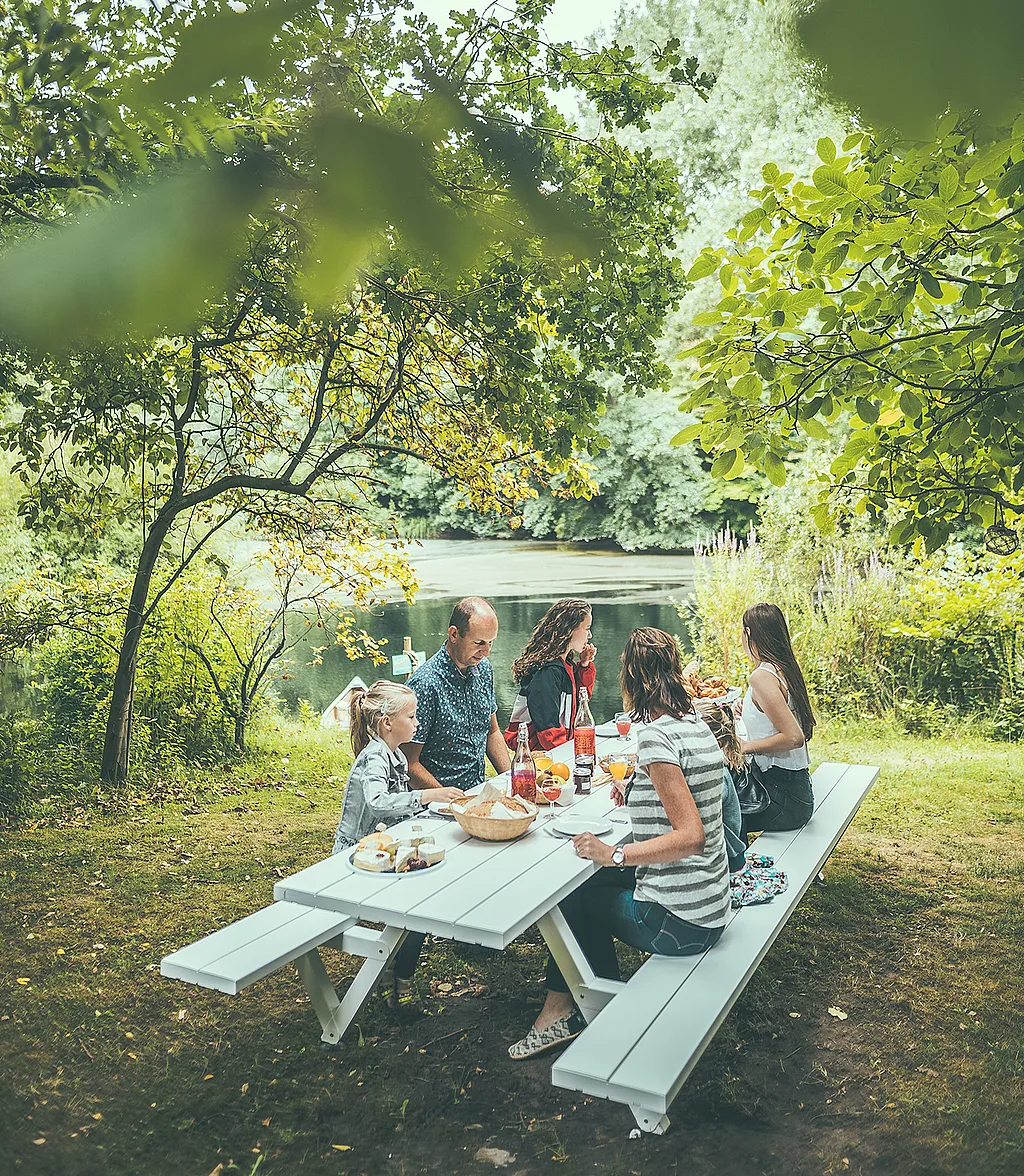 Une famille de cinq personnes déjeune ensemble à une table de pique-nique Marina gris clair, au bord d'un lac, sous des arbres feuillus, à l'hôtel éphémère The Pop-Up Hotel.