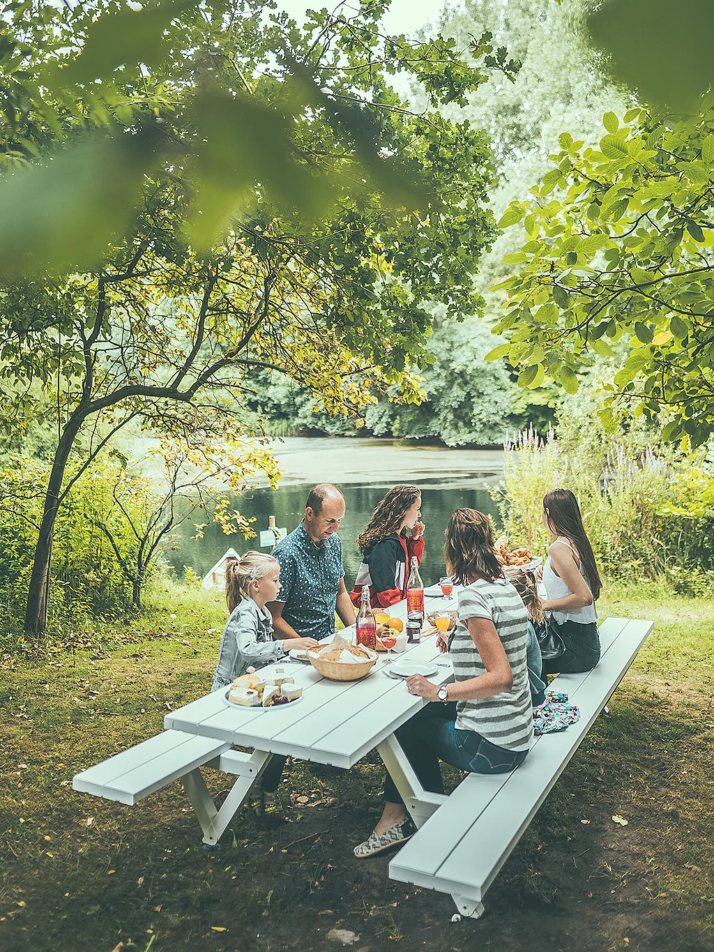 Eine fünfköpfige Familie isst gemeinsam an einem hellgrauen Picknicktisch am Seeufer, unter schattigen Bäumen im Pop-Up Hotel.