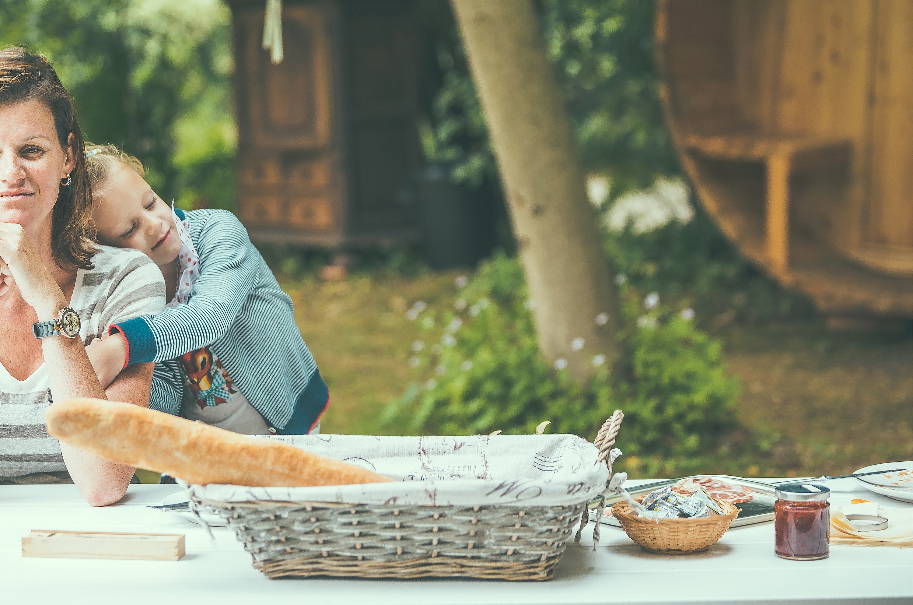 “A woman and child sit at a Marina picnic table with breakfast food and bread baskets, with wooden glamping cabins softly blurred in the background.