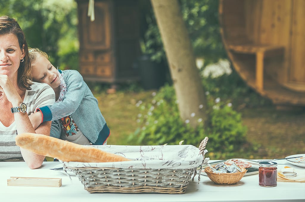 “A woman and child sit at a Marina picnic table with breakfast food and bread baskets, with wooden glamping cabins softly blurred in the background.