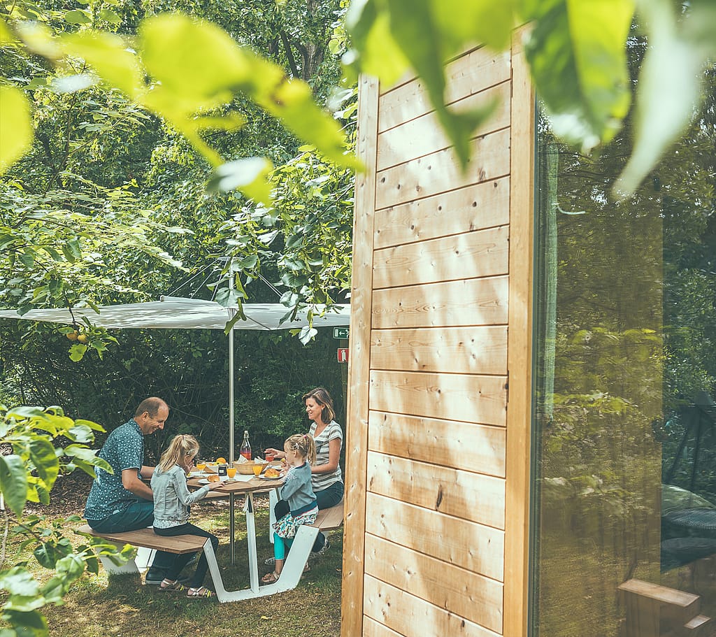 A family eats outdoors at an Anker picnic table under a white Inumbrina parasol beside a timber pop-up hotel cabin.